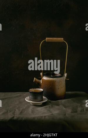 Craft handmade ceramic teapot kettle with cup of hot green tea for tea ceremony standing on linen tablecloth in dark room. Stock Photo
