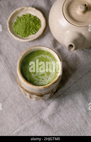 Traditional japanese hot green frothy tea matcha latte in handmade ceramic cup, powdered matcha, teapot and bamboo whisk standing on grey linen tablec Stock Photo