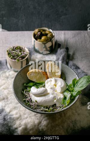 Traditional italian burrata knotted cheese salad in grey ceramic bowl on table. Sliced bread, olives, green sprouts around. Healthy mediterranean dinn Stock Photo