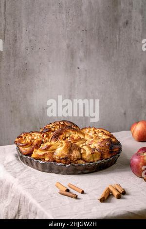 Sweet apple sticky cinnamon rolls buns in round baking tray standing with gardening apples and cinnamon sticks on grey linen table cloth. Traditional Stock Photo