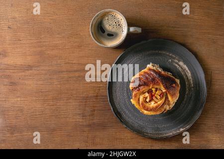 Sweet apple sticky cinnamon roll bun on balck plate with coffee mug standing on wooden background. Coffee break with bakery. Flat lay, copy space Stock Photo