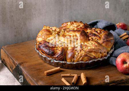 Sweet apple sticky cinnamon rolls buns in round baking tray standing with gardening apples and cinnamon sticks on wooden table. Traditional home baker Stock Photo