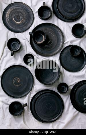 Set of empty black handcrafted ceramic plates, bowls and cups on white linen table cloth as background. Flat lay. Stock Photo