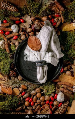 Empty black ceramic plate with cloth napkin over ambiance magic autumnal forest background. Autumn leaves, moss, fir cones, snail shell as surface. Au Stock Photo