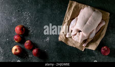 Raw organic uncooked young whole duck on crumpled paper with salt and gardening red apples around over black texture background. Top view, copy space. Stock Photo