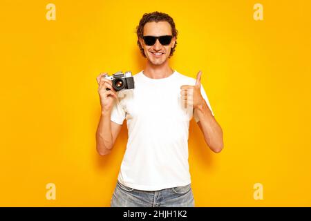 male photographer with retro camera in his hand gives thumbs up dressed in casual clothes sunglasses Stock Photo