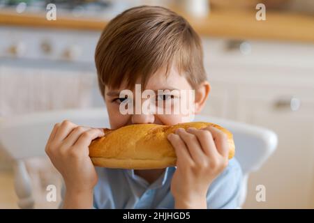 Happy handsome young teenage boy holding and eating freshly baked bread. A hungry boy is biting a big bread, in the kitchen at home. A child with handmade wheat flour bread looks into the camera. Stock Photo