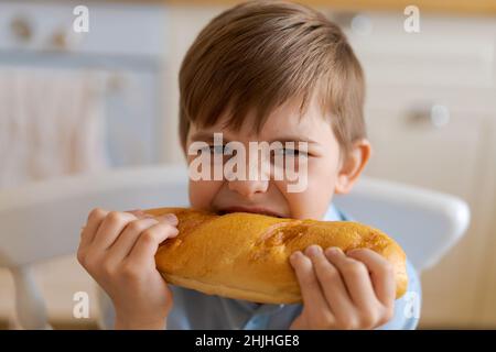 Happy handsome young teenage boy holding and eating freshly baked bread. A hungry boy is biting a big bread, in the kitchen at home. A child with handmade wheat flour bread looks into the camera. Stock Photo