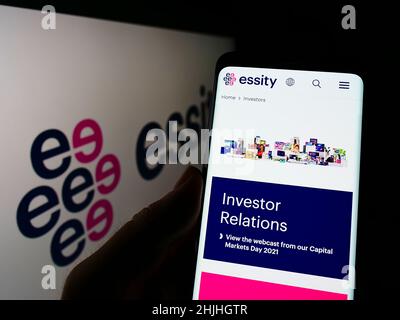 Person holding cellphone with webpage of Swedish consumer goods company Essity AB on screen in front of logo. Focus on center of phone display. Stock Photo