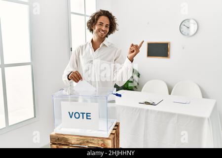 Young hispanic man voting putting envelop in ballot box with a big smile on face, pointing with hand finger to the side looking at the camera. Stock Photo