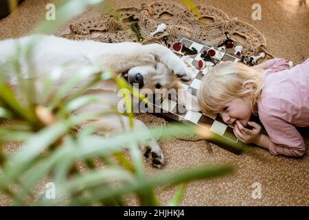 girl with a dog lie on the floor and have fun playing together. Golden Retriever puppy and little girl resting after a game of chess Stock Photo