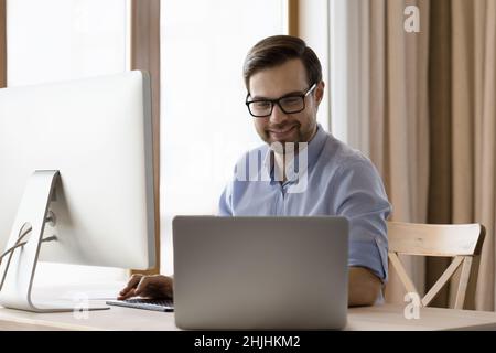 Happy smart millennial businessman in eyeglasses working on different gadgets. Stock Photo