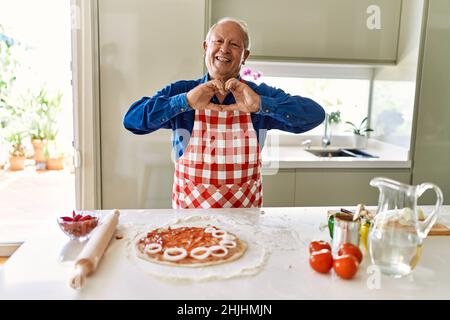 Senior man with grey hair cooking pizza at home kitchen smiling in love showing heart symbol and shape with hands. romantic concept. Stock Photo