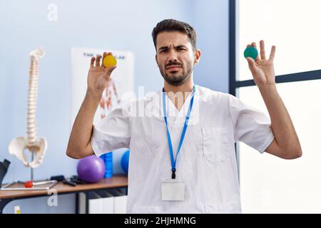 Young hispanic physiotherapist man holding strength balls to train hand muscles clueless and confused expression. doubt concept. Stock Photo