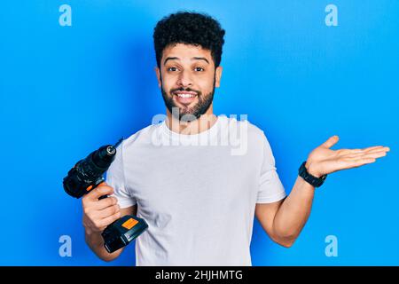 Young arab man with beard using drill celebrating achievement with happy smile and winner expression with raised hand Stock Photo