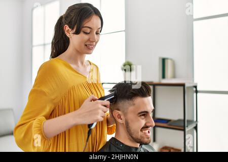 Young hispanic hairdresser woman cutting man's hair using electric razor machine at beauty center. Stock Photo