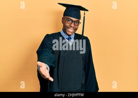 Young african american man wearing graduation cap and ceremony robe smiling cheerful offering palm hand giving assistance and acceptance. Stock Photo