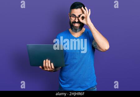 Hispanic man with beard working using computer laptop smiling happy doing ok sign with hand on eye looking through fingers Stock Photo