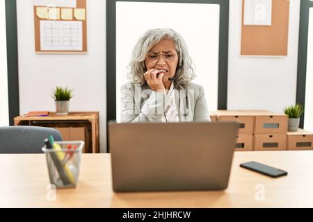 Middle age businesswoman sitting on desk working using laptop at office looking stressed and nervous with hands on mouth biting nails. anxiety problem Stock Photo