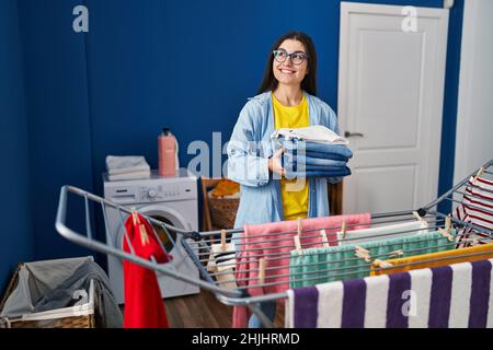 Young hispanic woman holding folded jeans hanging clothes on clothesline at laundry room Stock Photo