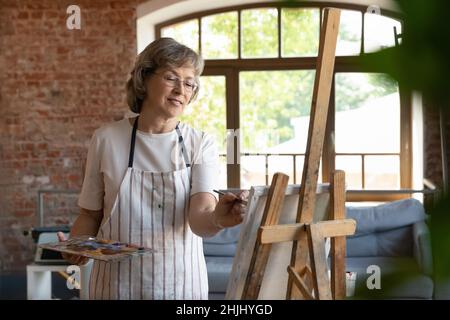 Happy senior retired lady drawing in paints on canvas Stock Photo