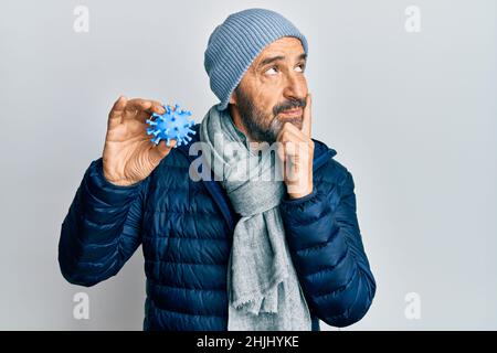 Middle age hispanic man wearing winter style holding virus toy serious face thinking about question with hand on chin, thoughtful about confusing idea Stock Photo
