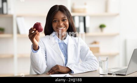 Pretty african american female nutritionist sitting in front of laptop and holding red apple, working in clinic Stock Photo