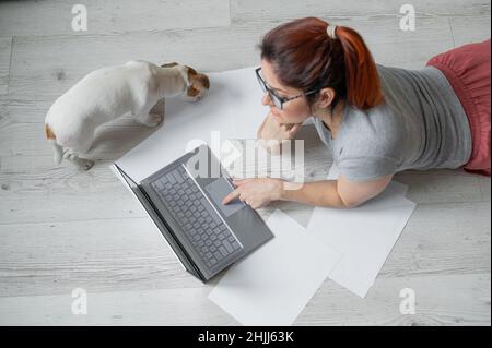 A woman lies on her stomach on the floor and is typing on a laptop. Girl freelancer works remotely from home with a puppy. Jack Russell Terrier next Stock Photo