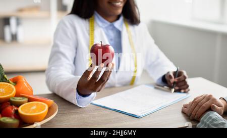 Healthy food concept. African american dietologist holding apple and consulting female patient in clinic Stock Photo