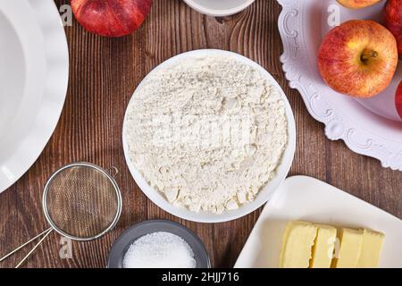 Bowl of flour surrounded by other pie crust ingredients like butter and sugar on wooden background Stock Photo