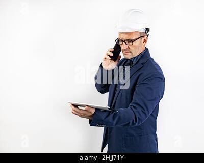 A male engineer in a white hard helmet is talking on a smartphone. Middle-aged man in a blue jacket with a tablet computer. Isolated portrait on a whi Stock Photo