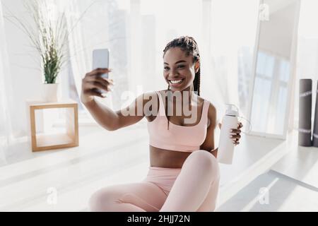 Charming young African American woman with bottle of water taking selfie during home workout Stock Photo