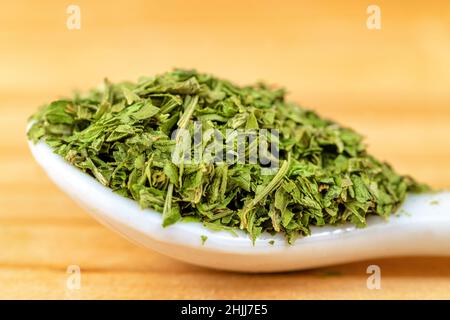 Closeup ceramic spoon with heap of dried green herbs placed on wooden table in kitchen Stock Photo
