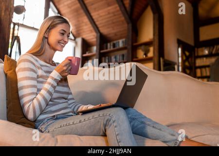 Smiling woman using laptop at home, drinking coffee Stock Photo