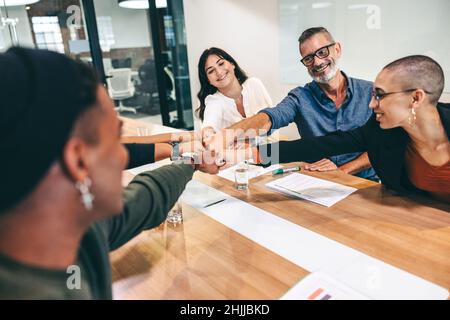 Successful group of businesspeople bringing their fists together while sitting in a boardroom. Diverse group of colleagues smiling cheerfully during a Stock Photo