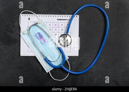 Paper calendar with date FEBRUARY 4, medical mask, stethoscope and awareness ribbon on dark background. World Cancer Day Stock Photo