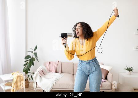 Happy young black lady with hair dryer as microphone singing and dancing at home, copy space Stock Photo