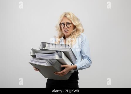 Stressed office worker overloaded with papers, holding folders of documents, standing on light grey studio background Stock Photo