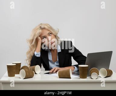 Exhausted woman office employee sitting at workplace surrounded by coffee cups, feeling tired of overtime work Stock Photo