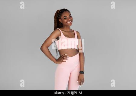 Portrait of lovely smiling young black female in comfy sports outfit posing and smiling at camera on grey studio background Stock Photo