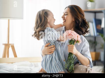 Happy day! Child daughter is congratulating mother and giving her flowers. Mum and girl smiling and hugging. Family holiday and togetherness. Stock Photo