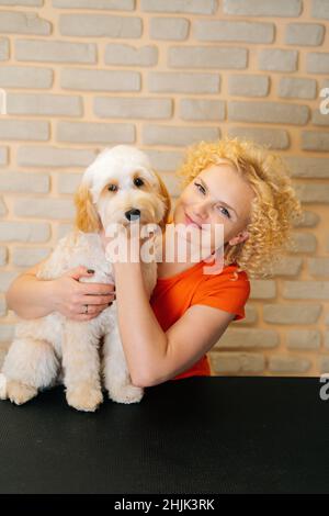 Vertical portrait of smiling female groomer holding curly Labradoodle dog after haircutting at grooming salon. Stock Photo