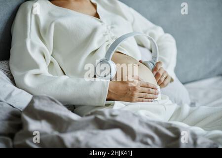 Close-up of pregnant woman putting on headphones on her belly and turning on the music while lying on the bed Stock Photo