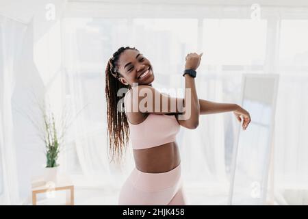 Portrait of attractive fit African American lady in sportswear exercising at home, stretching her arm and smiling at camera Stock Photo