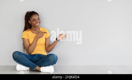 Cheery young black woman sitting cross legged on floor, pointing aside at free space against grey wall, banner Stock Photo