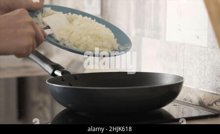 Side view of woman hands putting cut onion into pan to fry. Close up of woman putting fresh onion from the glass plate into the pan in the kitchen. Stock Photo