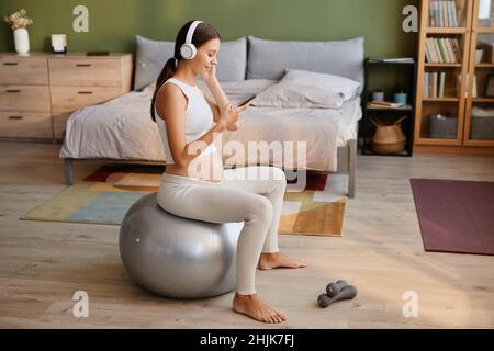 Young pregnant woman in headphones using mobile phone to listen to music while exercising on fitness ball in the bedroom Stock Photo