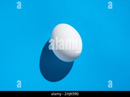 A white egg on a blue background with a hard shadow. The concept of Easter, minimalistic food. Stock Photo