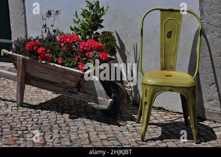 Street decoration-red azalea and violet flowers-wooden wheelbarrow-lemon yellow chair. Tavira-Portugal-088 Stock Photo