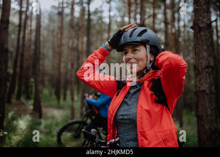 Senior woman biker putting on cycling helmet outdoors in forest in autumn day. Stock Photo